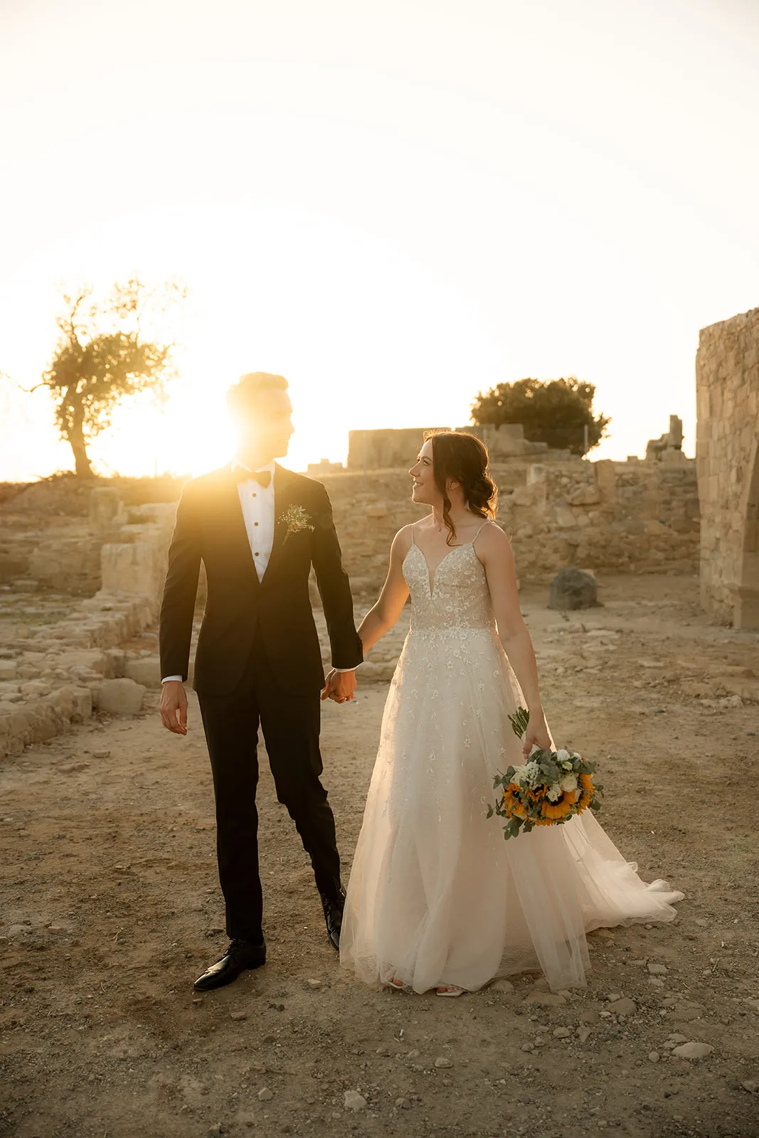 Bride and groom walking hand in hand, backlit by a low sunburst among stone ruins at sunset in Cyprus