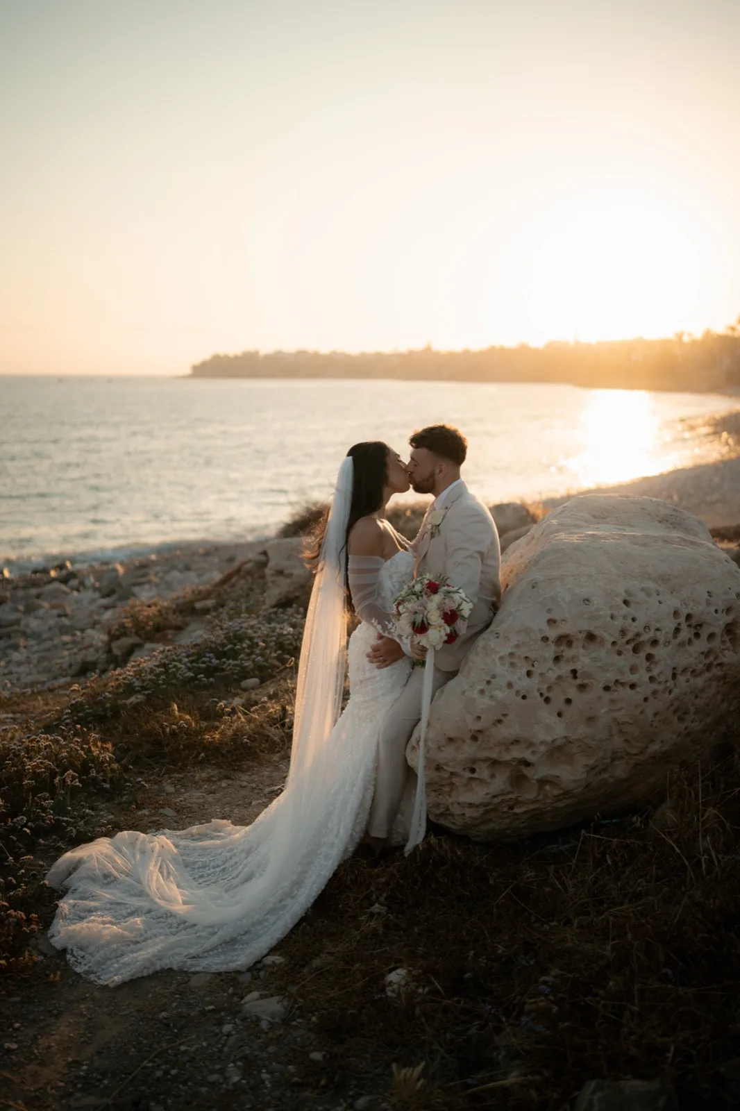 Bride and groom kissing on a limestone rock as the sun sets over the sea at Coral Residences, Paphos, Cyprus