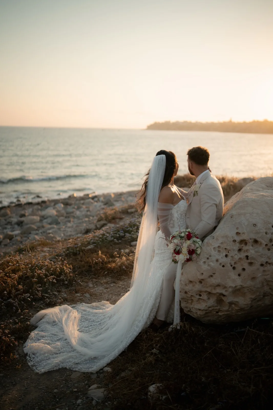 Bride and groom at golden hour, resting against a weathered limestone rock on the beach with the veil and dress train trailing across the sand at Coral Residences, Paphos, Cyprus