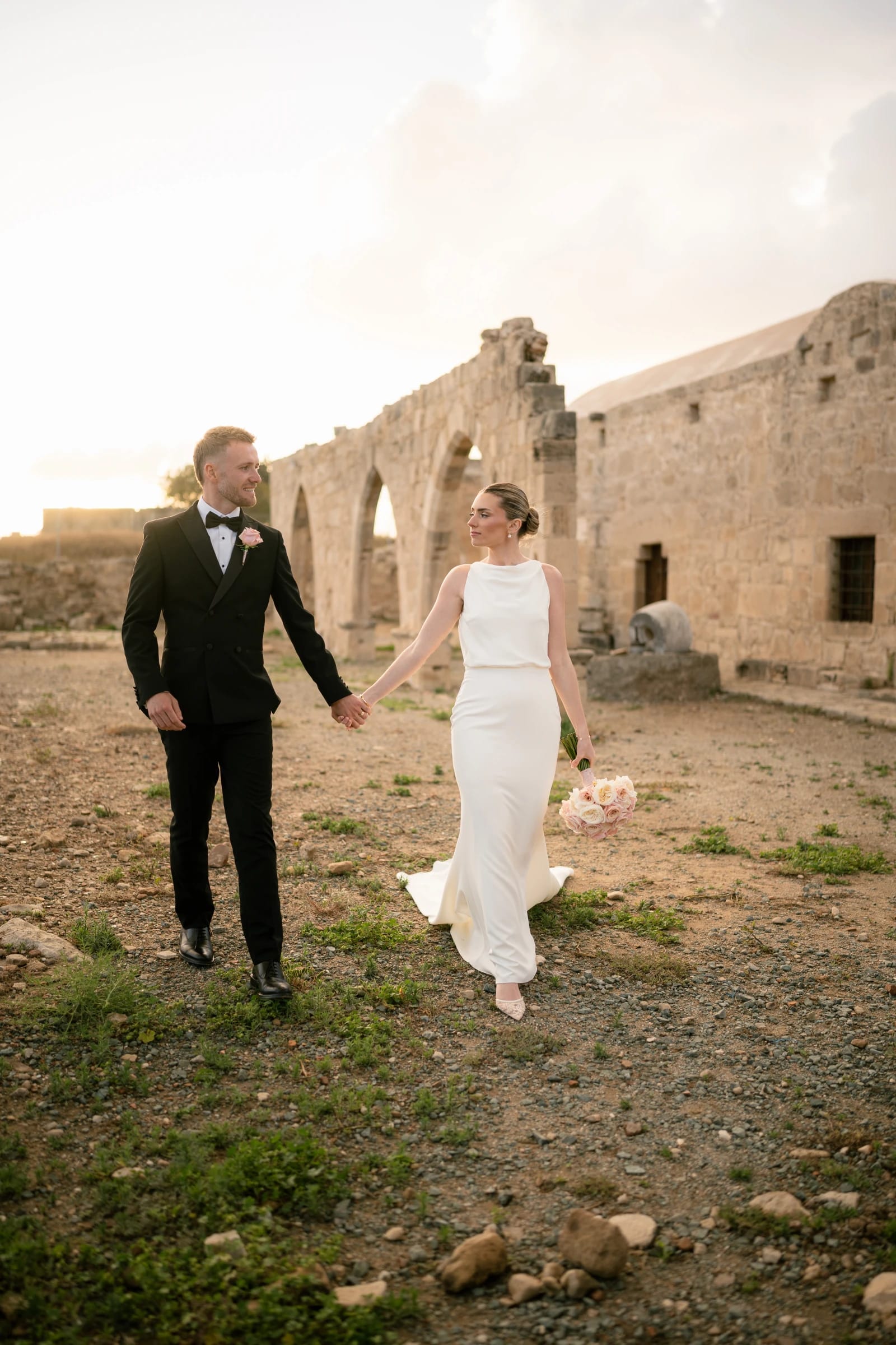 Bride and groom walking hand in hand through stone ruins at sunset at Liopetro, Kouklia, Paphos