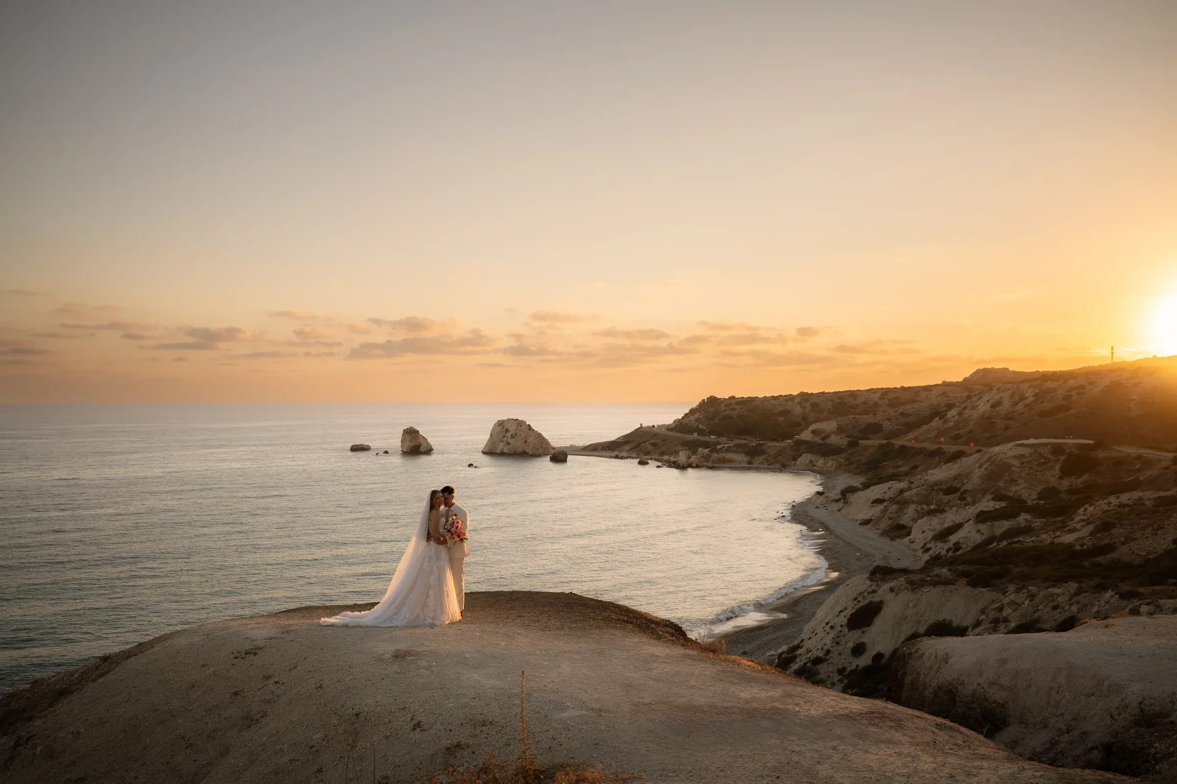 Frank and Franziska embracing on the headland above the bay at Aphrodite's Rock as the sun sets over the sea, after their Liopetro wedding near Paphos, Cyprus.