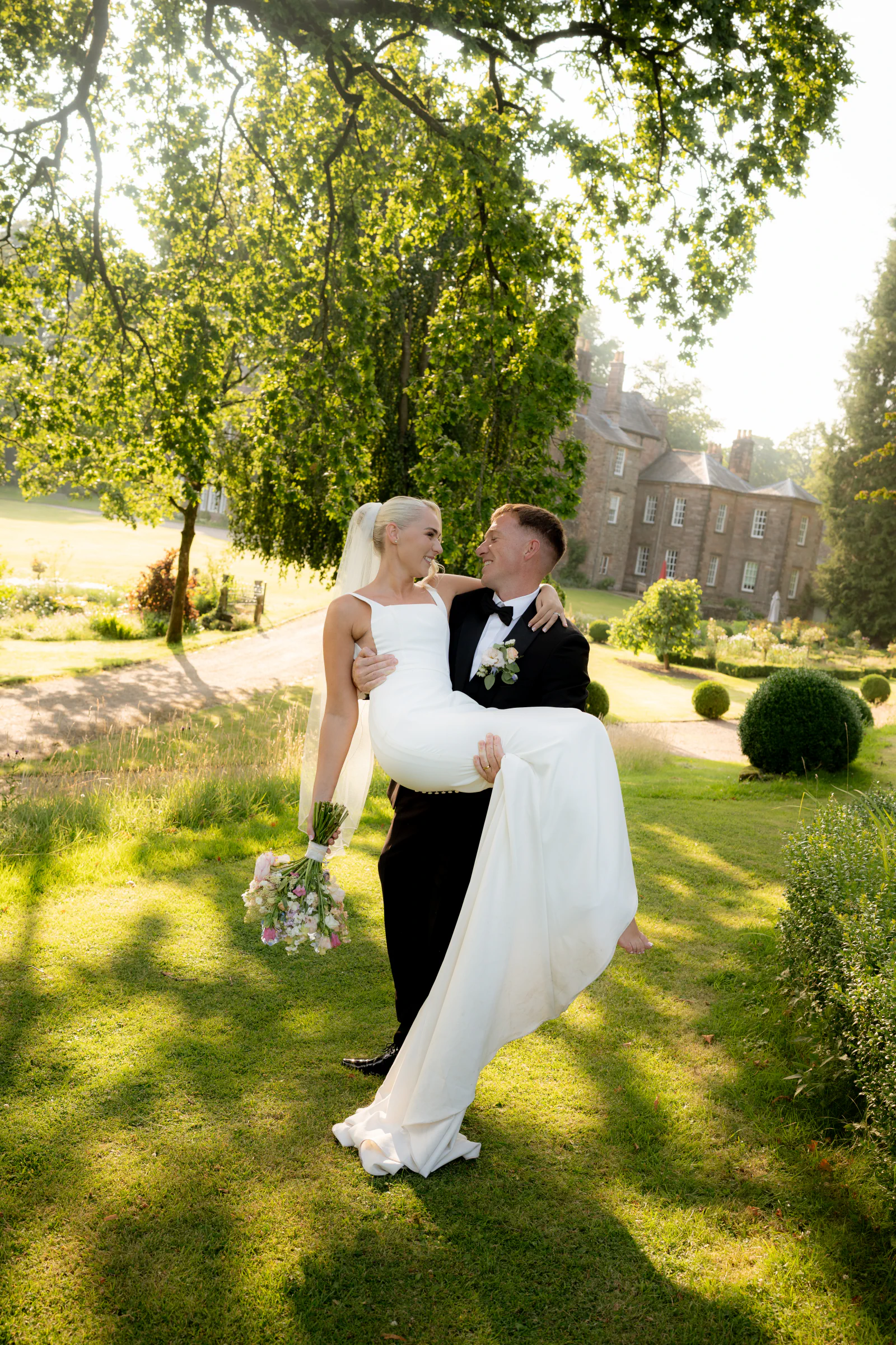 Josh carrying Ellie through the sunlit gardens with Browsholme Hall behind them.