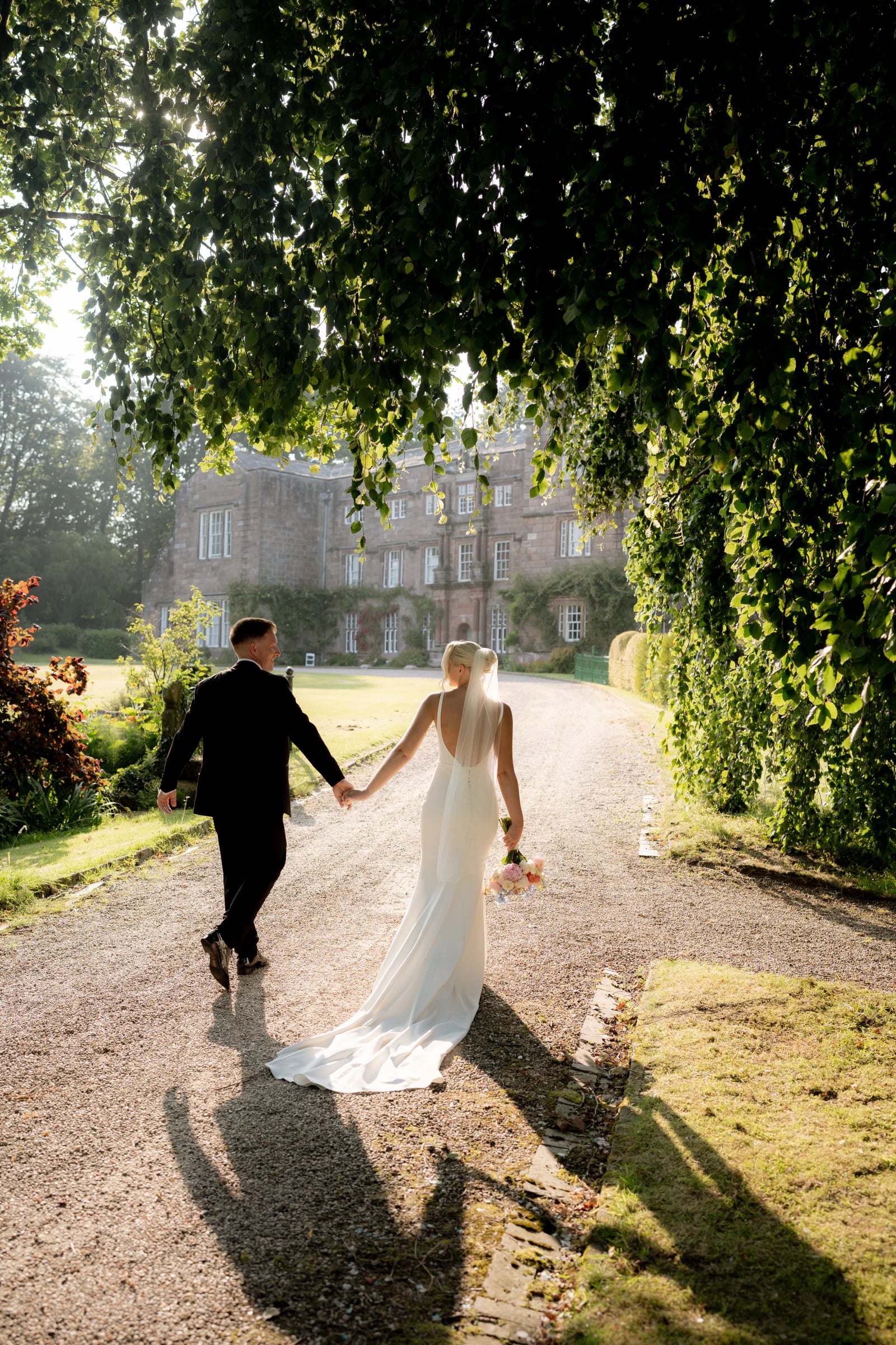 Josh and Ellie walking hand in hand towards Browsholme Hall beneath the trees in the evening light.