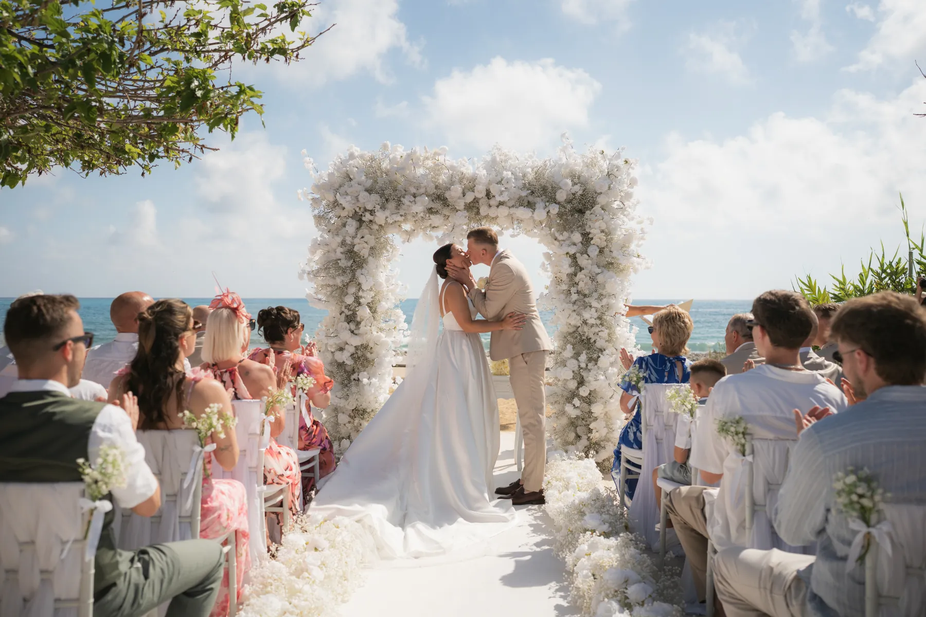 Sian and Aaron sharing their first kiss beneath a white floral arch with the sea behind them at Coral Residences, Paphos, Cyprus.