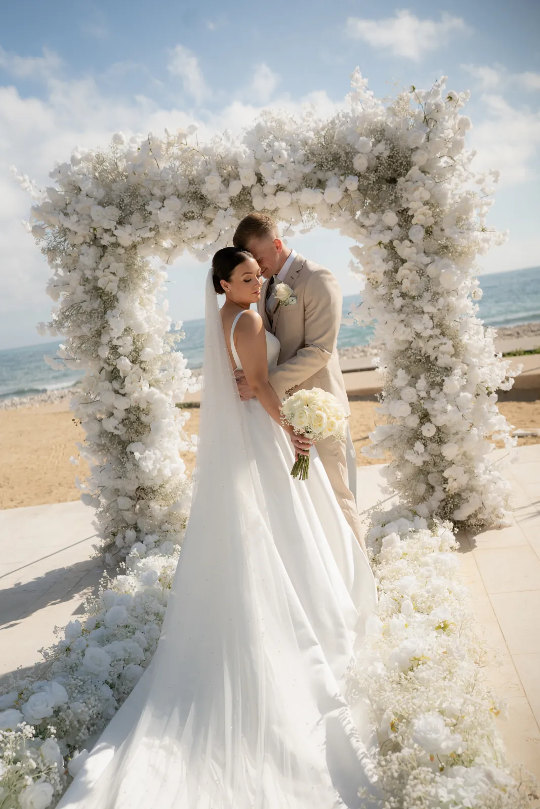 Bride and groom embracing under a white floral arch with the sea behind at Coral Residences, Paphos, Cyprus